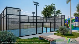 Vista de una moderna cancha de Pickleball al aire libre en un complejo residencial. La cancha tiene una superficie de color azul claro y está cercada por una estructura de malla metálica de color negro. Se aprecian postes de iluminación alta (con faros LED) para jugar de noche. En primer plano hay una banca de madera sobre un camino pavimentado y césped bien cuidado. Al fondo se distingue una zona de juegos infantiles con colores brillantes. En la esquina se lee 'PICKLEBALL VITA RESIDENCIAL' con información de contacto.